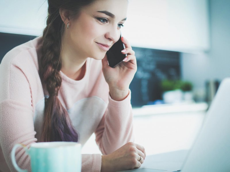 Young woman sits at the kitchen table using a laptop and talking on a cell phone